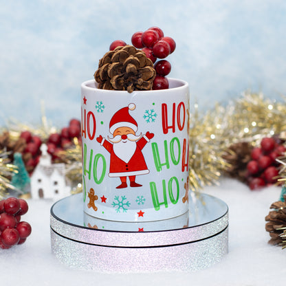 A white ceramic mug with a festive 'Ho Ho Ho' Santa Claus design, styled on a round mirrored pedestal with pinecones and red berries against a soft blue holiday backdrop.