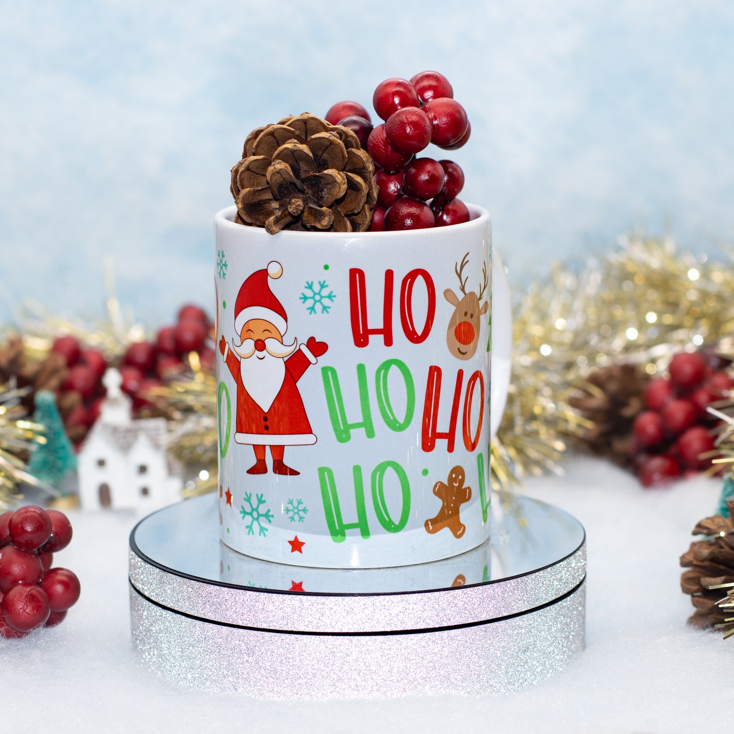 A white ceramic mug with a festive 'Ho Ho Ho' Christmas print, styled with a pinecone and red berries on top, sitting on a round silver glitter display stand.