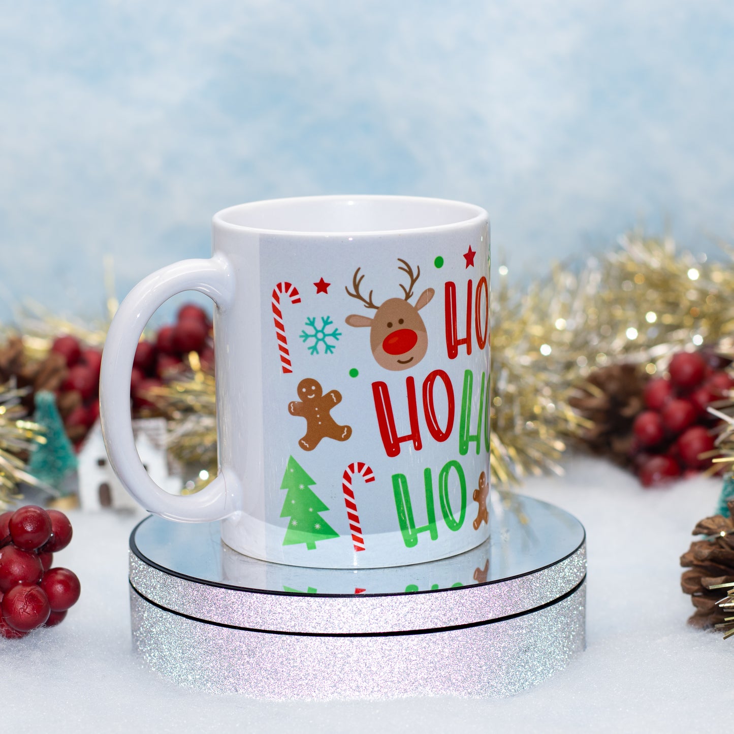 A white ceramic mug with a festive 'Ho Ho Ho' holiday print featuring a reindeer, gingerbread man, candy canes, and a Christmas tree, displayed on a round silver glitter pedestal with holiday decorations in the background.