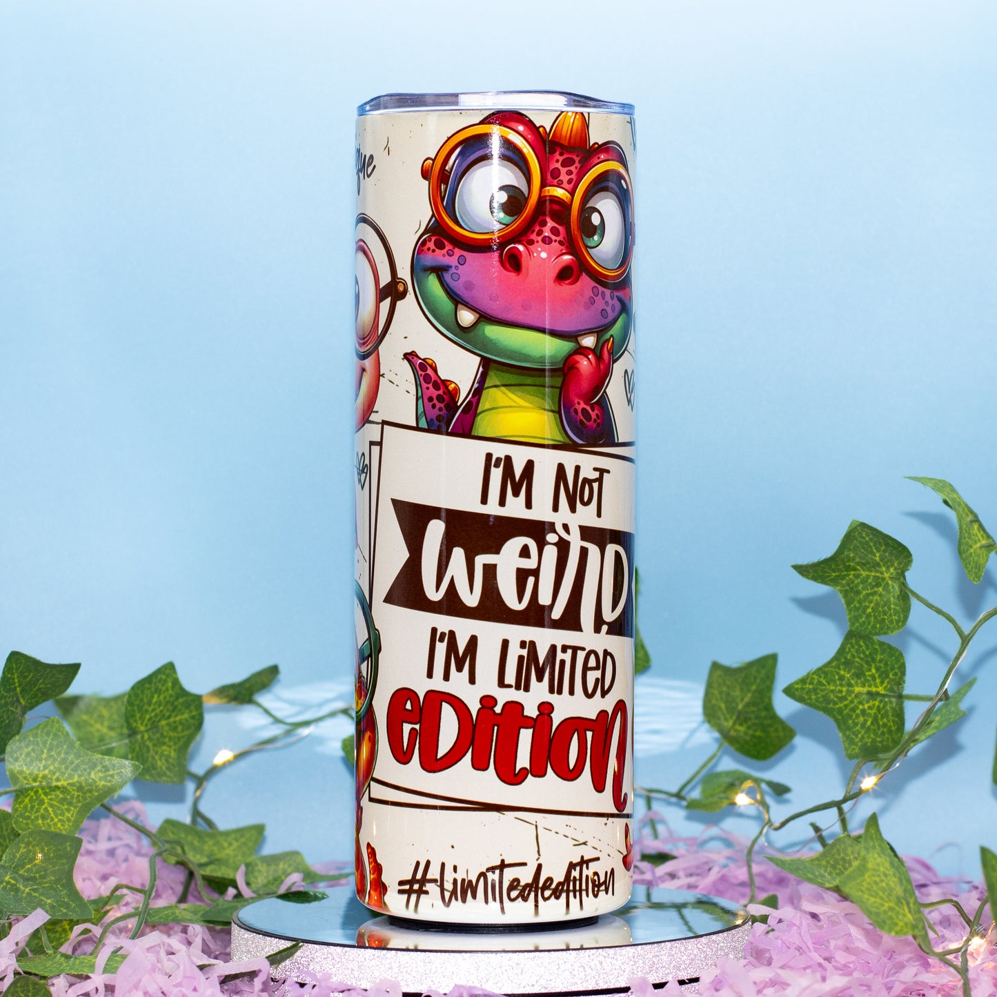 A cylindrical tumbler with a colorful cartoon dino graphic and the text 'I'M Not weird I'M Limited Edition' and '#limitededition', displayed on a mirrored stand with pink shredded paper and green ivy against a light blue background.
