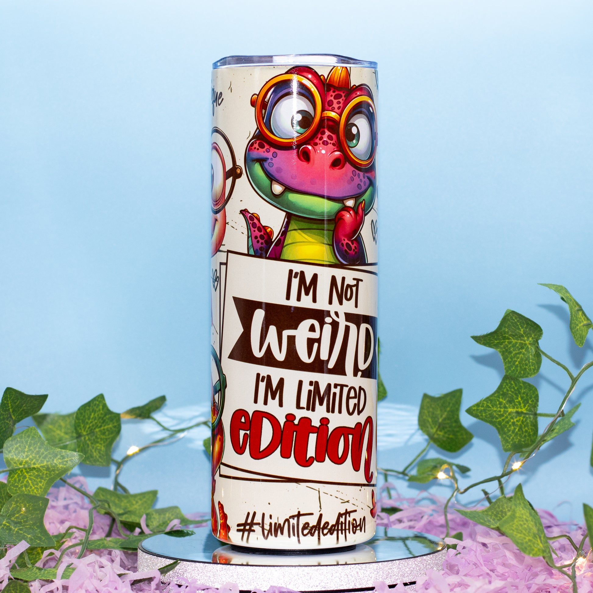 A cylindrical tumbler with a colorful cartoon dino graphic and the text 'I'M Not weird I'M Limited Edition' and '#limitededition', displayed on a mirrored stand with pink shredded paper and green ivy against a light blue background.