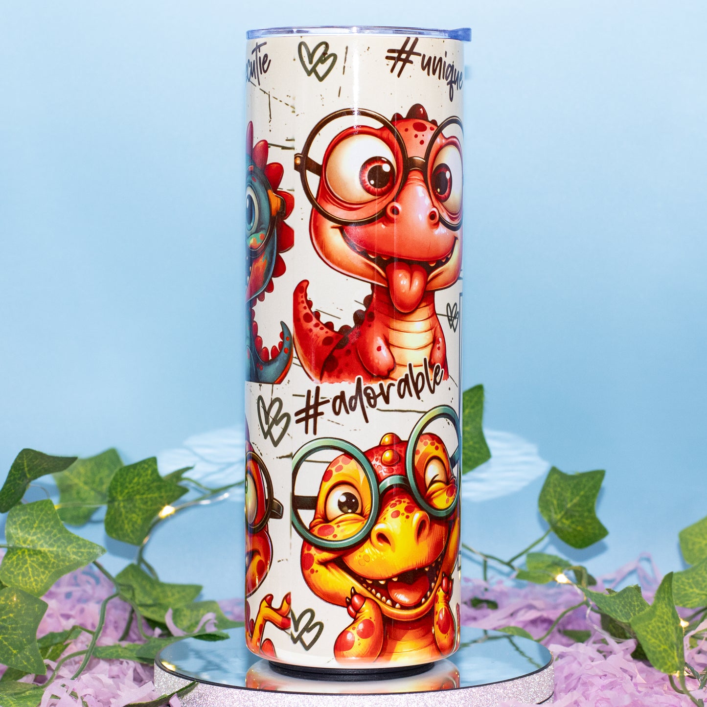 A tall insulated tumbler with a cute cartoon dinosaur wearing round glasses, shown standing upright on a reflective surface with decorative green leaves and purple flowers against a light blue background.