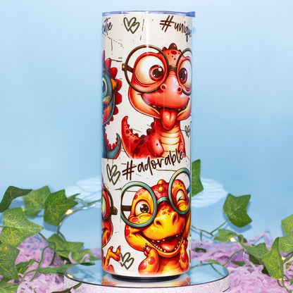 A tall insulated tumbler with a cute cartoon dinosaur wearing round glasses, shown standing upright on a reflective surface with decorative green leaves and purple flowers against a light blue background.