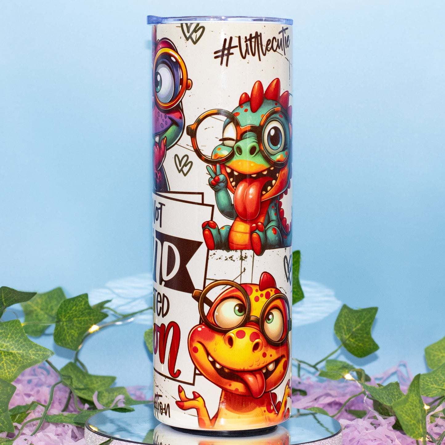 A tall cylindrical tumbler with a white background and colorful cartoon dinosaur illustrations, displayed on a reflective surface with decorative green leaves and purple flowers against a light blue background.