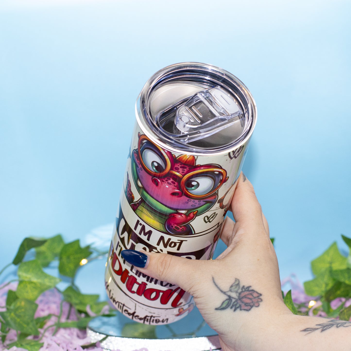 A hand holds a stainless steel tumbler with a clear flip straw lid. The tumbler features a white background with a colourful cartoon dino wearing glasses 