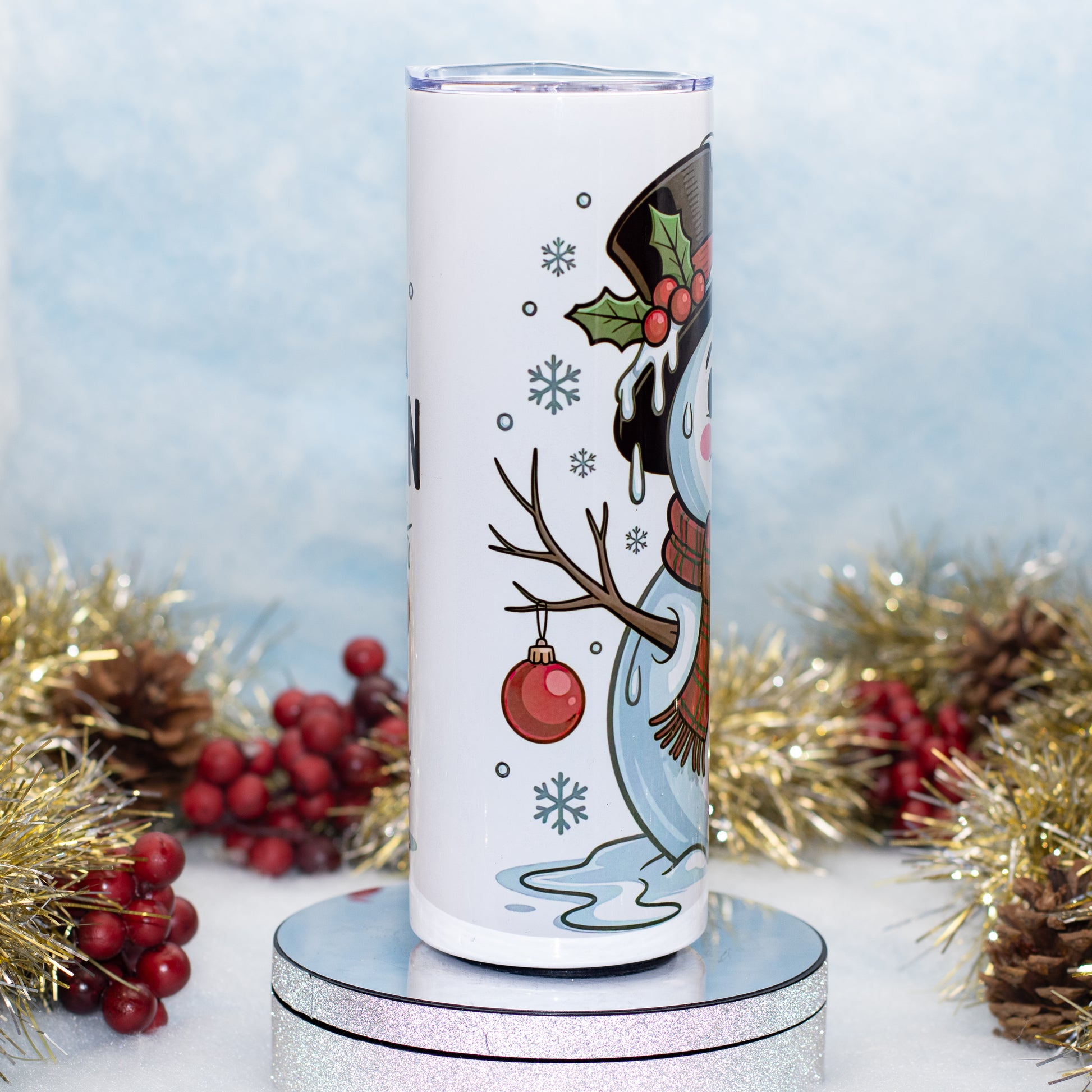 A white insulated tumbler with a festive Christmas snowman illustration, displayed on a round silver stand with holiday decorations in the background.