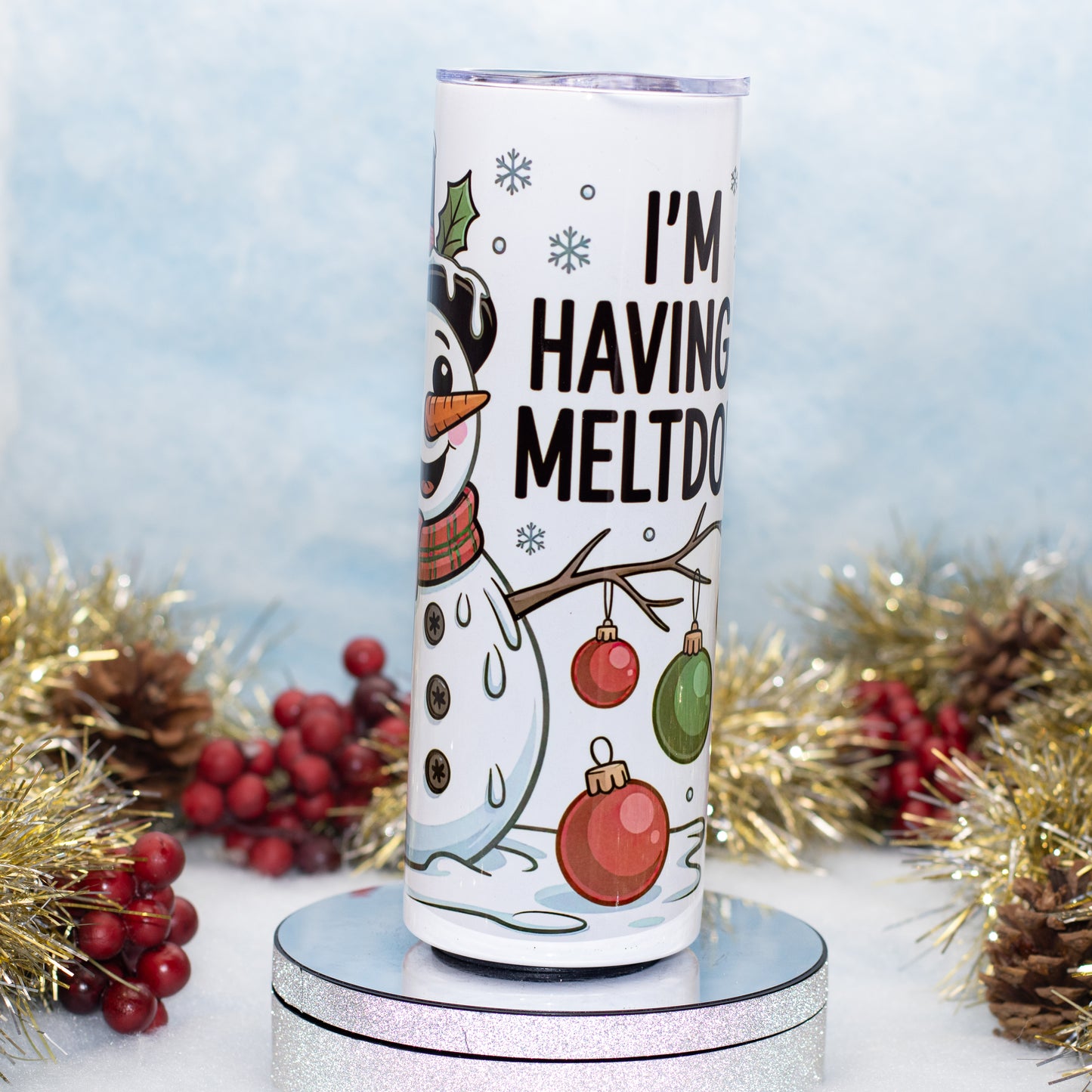 A white insulated tumbler with a holiday snowman illustration and the text 'I'M HAVING MELTDOWN' displayed on a round pedestal, surrounded by gold tinsel, pinecones, and red berries against a soft blue background.