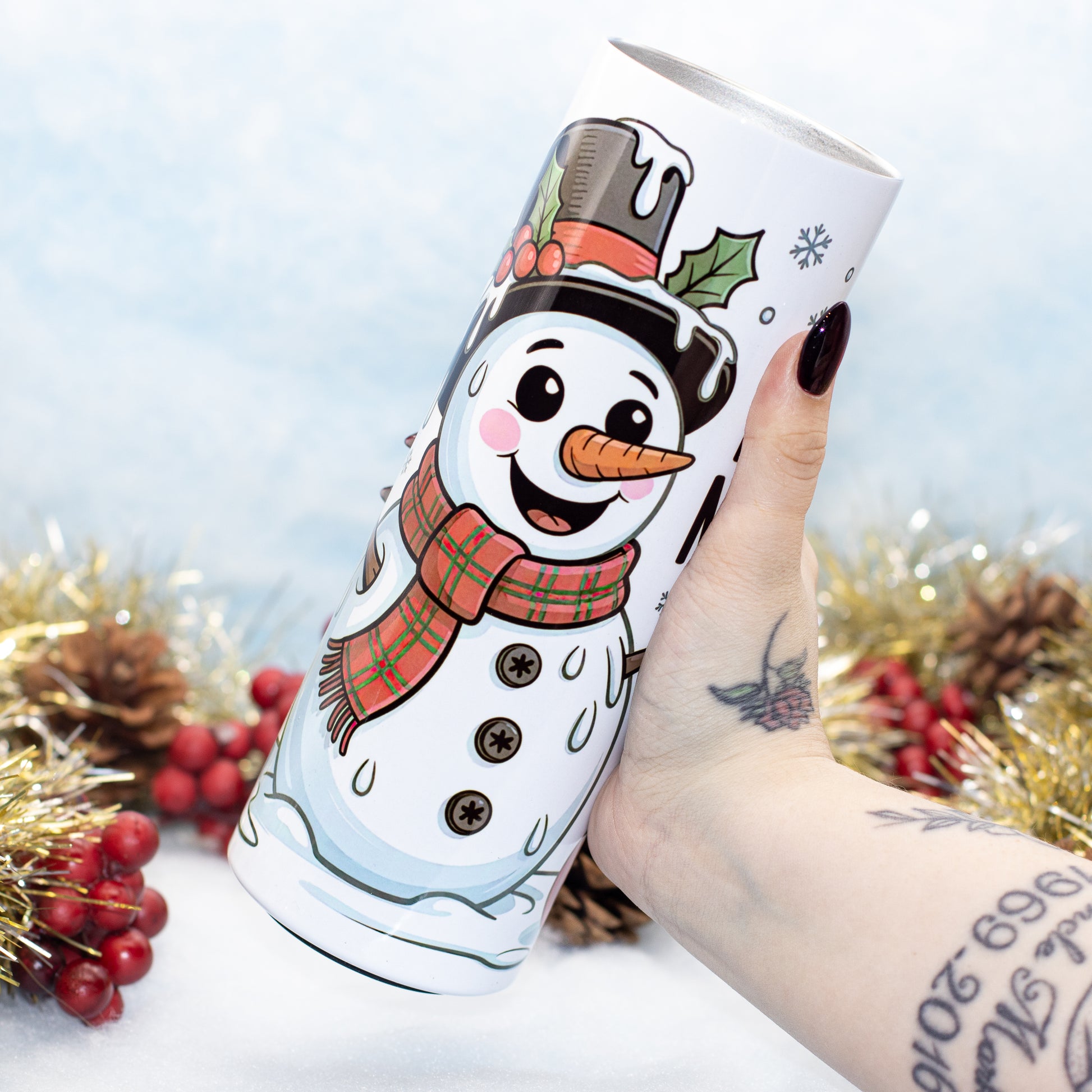 A hand holds a white insulated tumbler featuring a cartoon snowman wearing a black top hat with holly and a red plaid scarf. The background is a festive, snowy scene with gold tinsel, pinecones, and red berries.