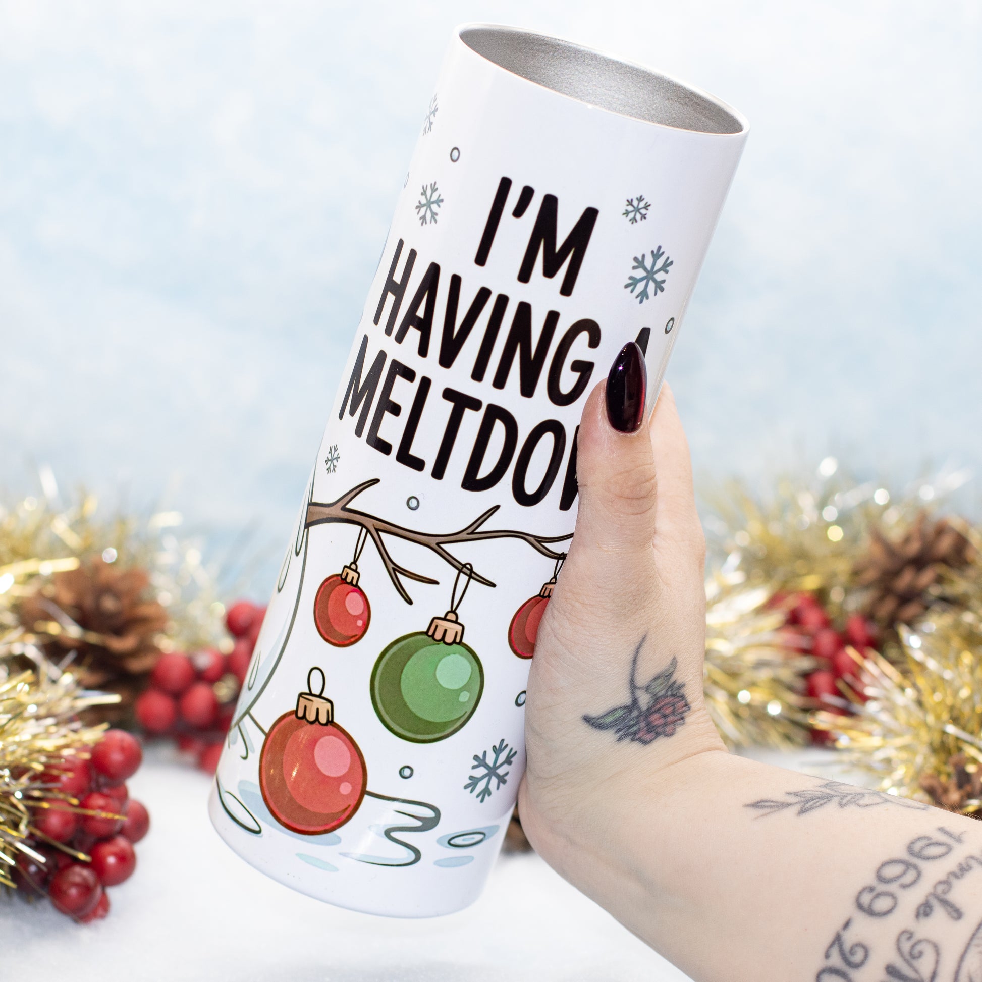 A hand holds a white insulated tumbler decorated with the text 'I'M HAVING MELTDOWN' and Christmas ornament illustrations. The background is decorated with gold tinsel, pinecones, and red berries against a light blue textured backdrop.