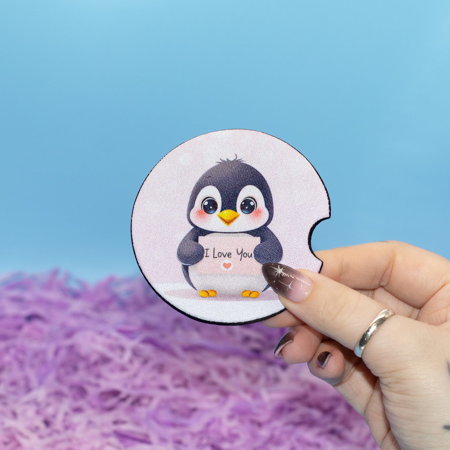 A hand hold a car coaster featuring a cute cartoon penguin holding a sign that says 'I Love You'. The background consists of a solid light blue upper section and a textured purple shredded paper filler at the bottom.