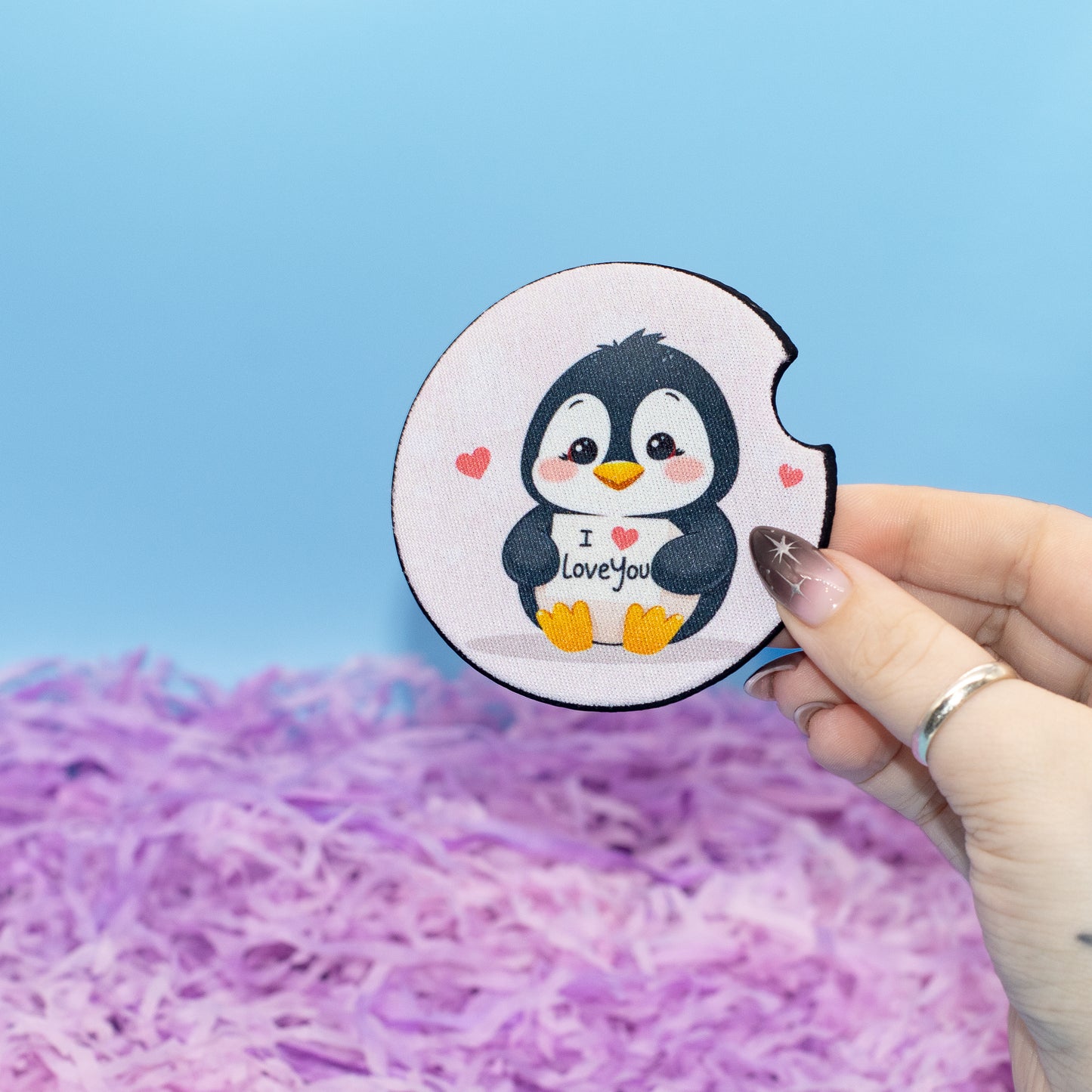A hand holds a round white coaster featuring a cute cartoon penguin with black and white feathers, yellow feet and beak, and small pink hearts. The penguin holds a sign that says 'I love you'.