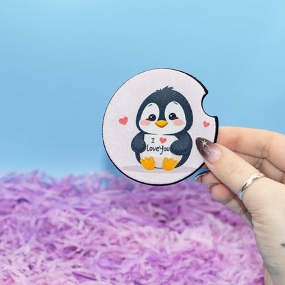 A hand holds a round white coaster featuring a cute cartoon penguin with black and white feathers, yellow feet and beak, and small pink hearts. The penguin holds a sign that says 'I love you'.