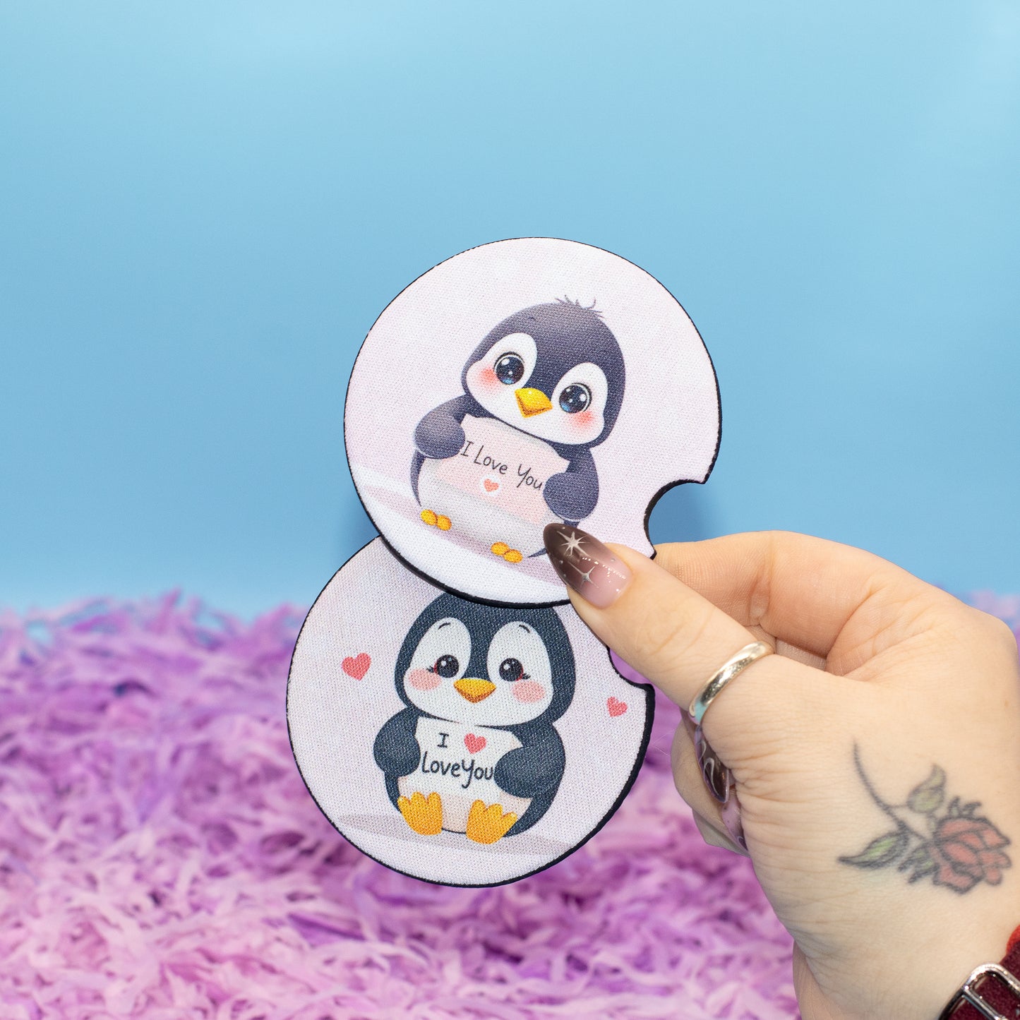 A hand holds two round car coasters with cartoon penguin illustrations against a blue background with purple shredded paper.