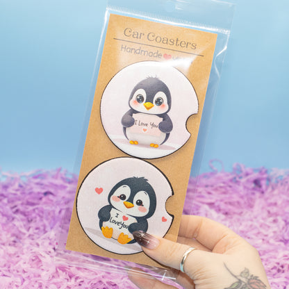 A hand holds a clear plastic bag containing two round car coasters with cute cartoon penguin designs on a kraft card. The background is a light blue wall with purple shredded paper filler.