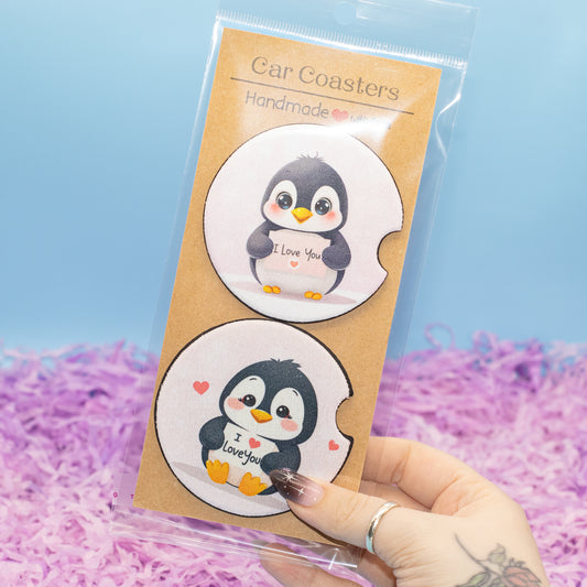A hand holds a clear plastic bag containing two round car coasters with cute cartoon penguin designs on a kraft card. The background is a light blue wall with purple shredded paper filler.
