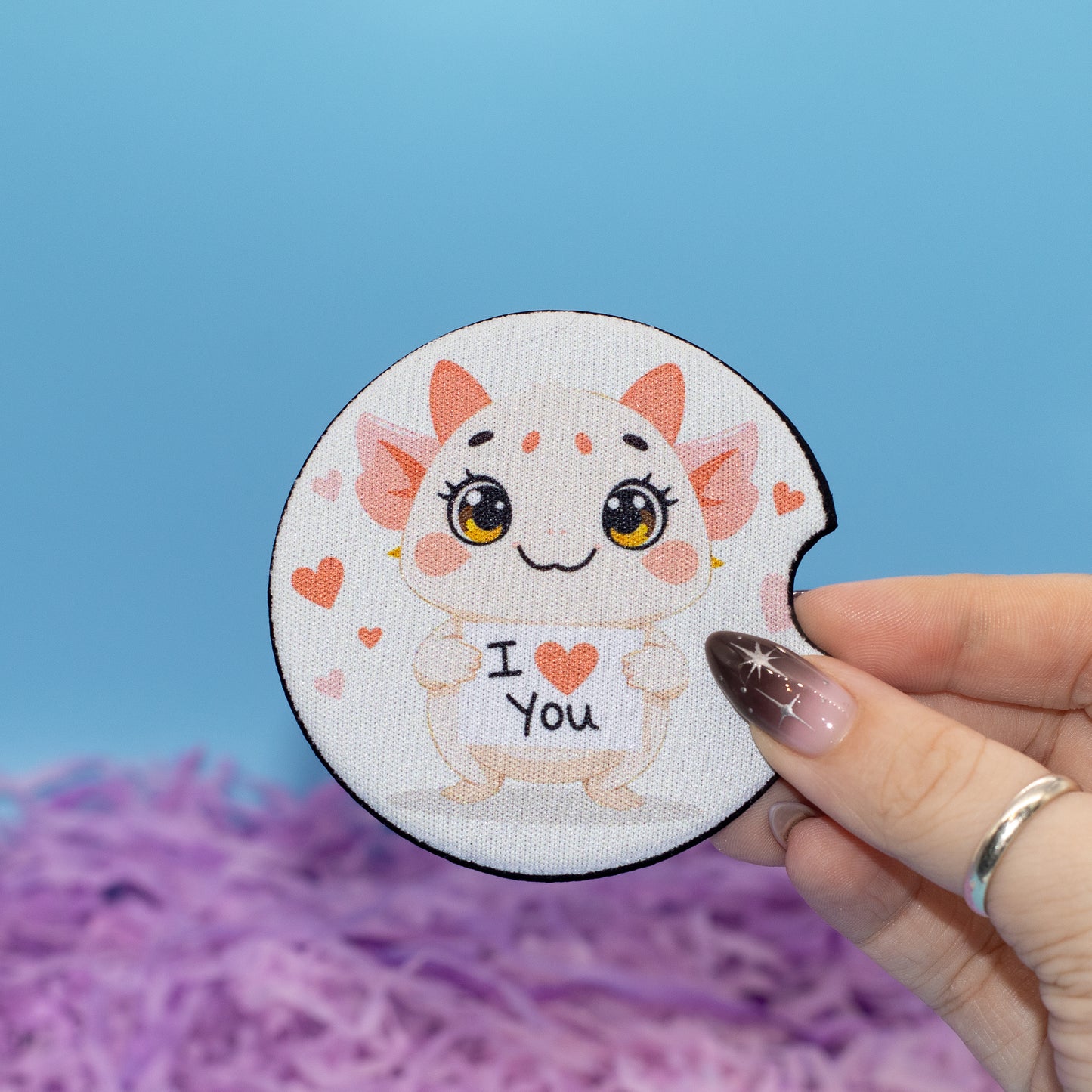 A hand holds car coaster featuring a cute cartoon creature with large eyes and the text 'I <heart> You' on a white background with a blue backdrop and purple shredded paper.