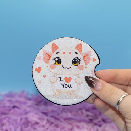 A hand holds car coaster featuring a cute cartoon creature with large eyes and the text 'I <heart> You' on a white background with a blue backdrop and purple shredded paper.
