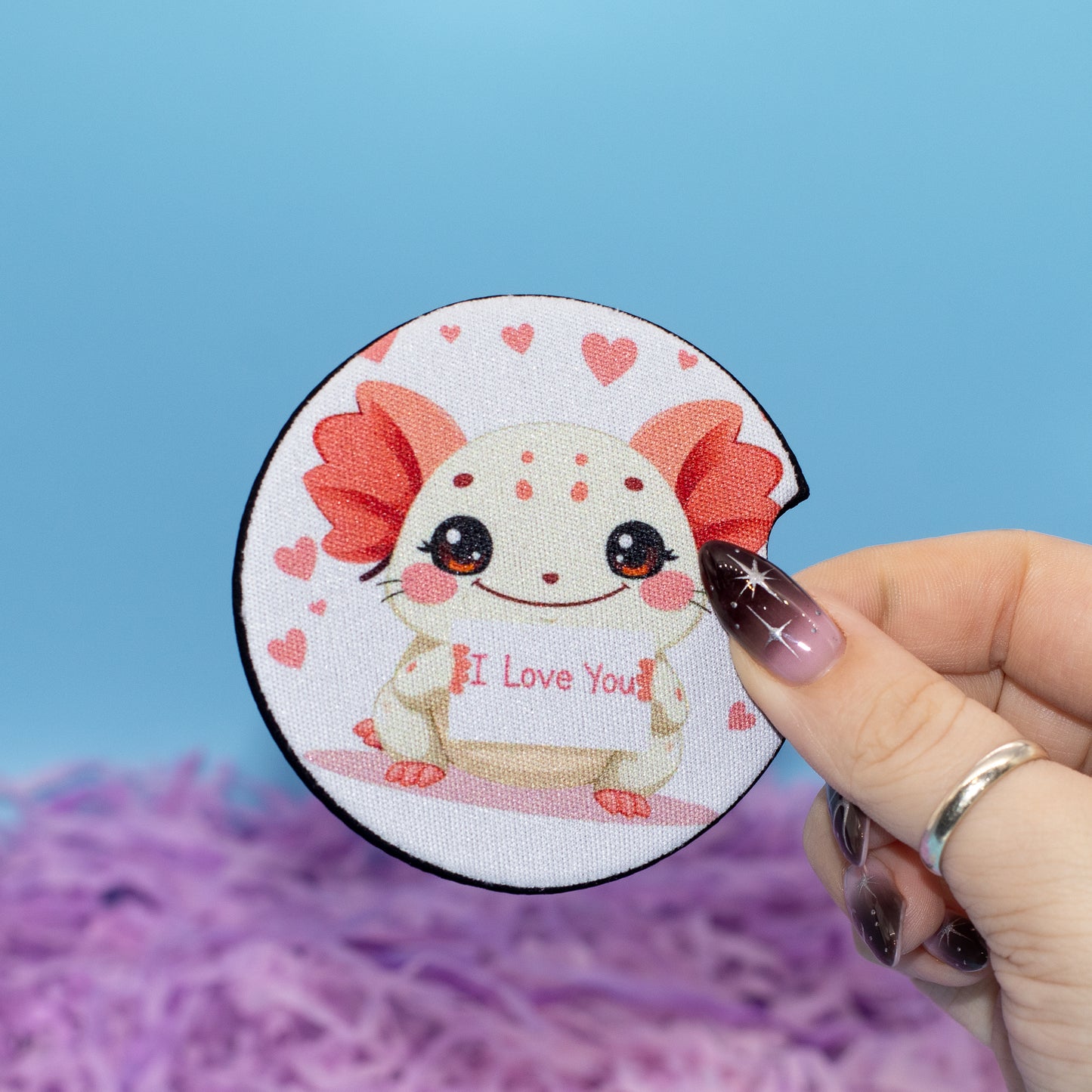 A hand hold a car coaster featuring a cute white axolotl with red gills and hearts, holding a sign that says 'I Love You'. The background is a bright blue sky with purple fluffy artificial grass.