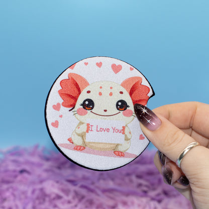 A hand hold a car coaster featuring a cute white axolotl with red gills and hearts, holding a sign that says 'I Love You'. The background is a bright blue sky with purple fluffy artificial grass.