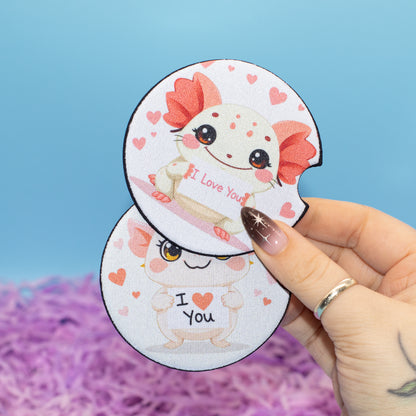 A hand holds two car coasters with cute axolotl illustrations with hearts and the text 'I Love You' and 'I <heart> You'. The background is a soft blue with purple fluffy filler.