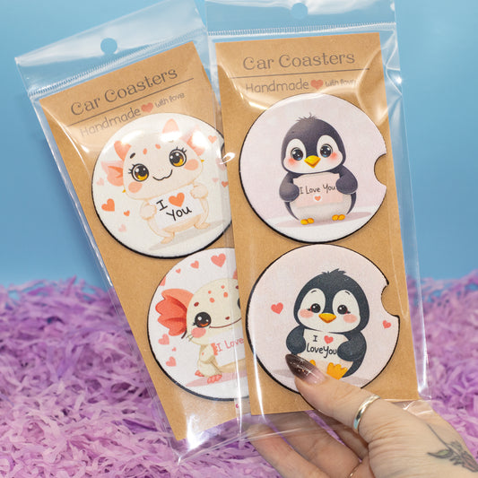 Two packaged sets of round car coasters with cute cartoon penguin and Axolotl illustrations, held by a hand over purple shredded paper against a blue background.