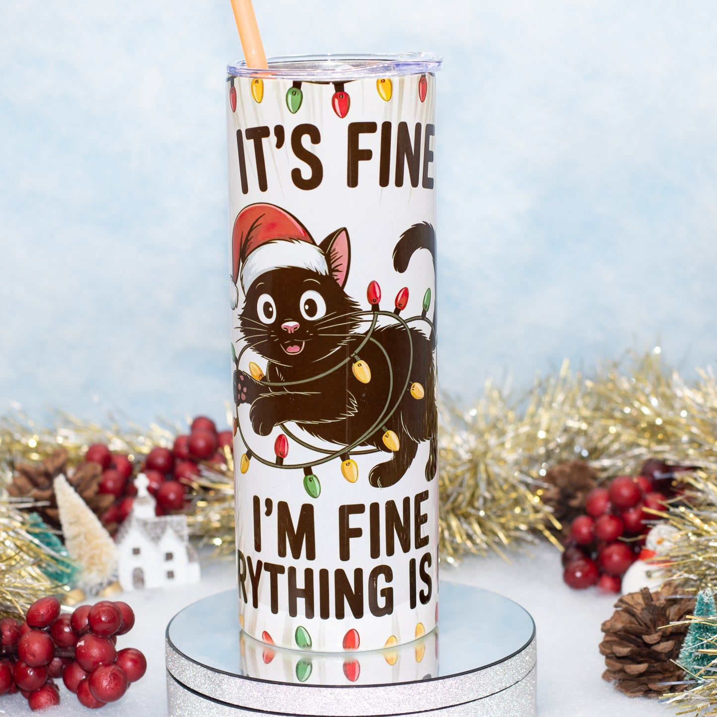 A white insulated tumbler with a clear lid and straw, printed with a cartoon black cat wearing a Santa hat and wrapped in holiday lights, with the text 'IT'S FINE' and 'I'M FINE' around it. The tumbler is shown on a silver pedestal with Christmas decorations in the background.