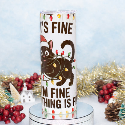 A white insulated tumbler with a holiday-themed design featuring a cartoon black cat wearing a Santa hat and wrapped in colouful Christmas lights, with the text 'IT'S FINE I'M FINE EVERYTHING IS FINE'. The tumbler is displayed on a silver pedestal with festive tinsel, pinecones, and red berries in the background.