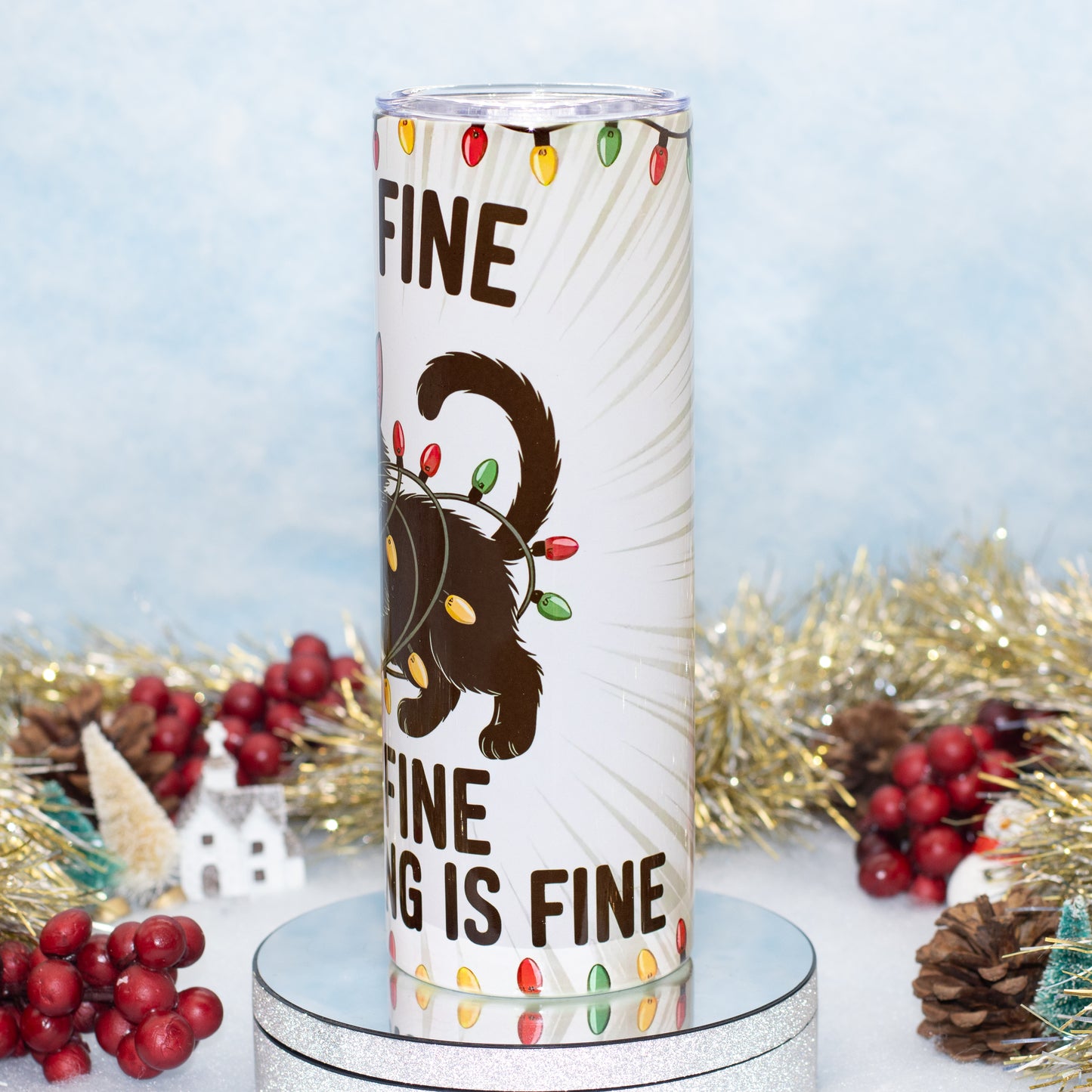A white cylindrical tumbler with a holiday design featuring a black cat wearing a string of multicolour Christmas lights, surrounded by festive tinsel, pinecones, and red berries on a light blue background.