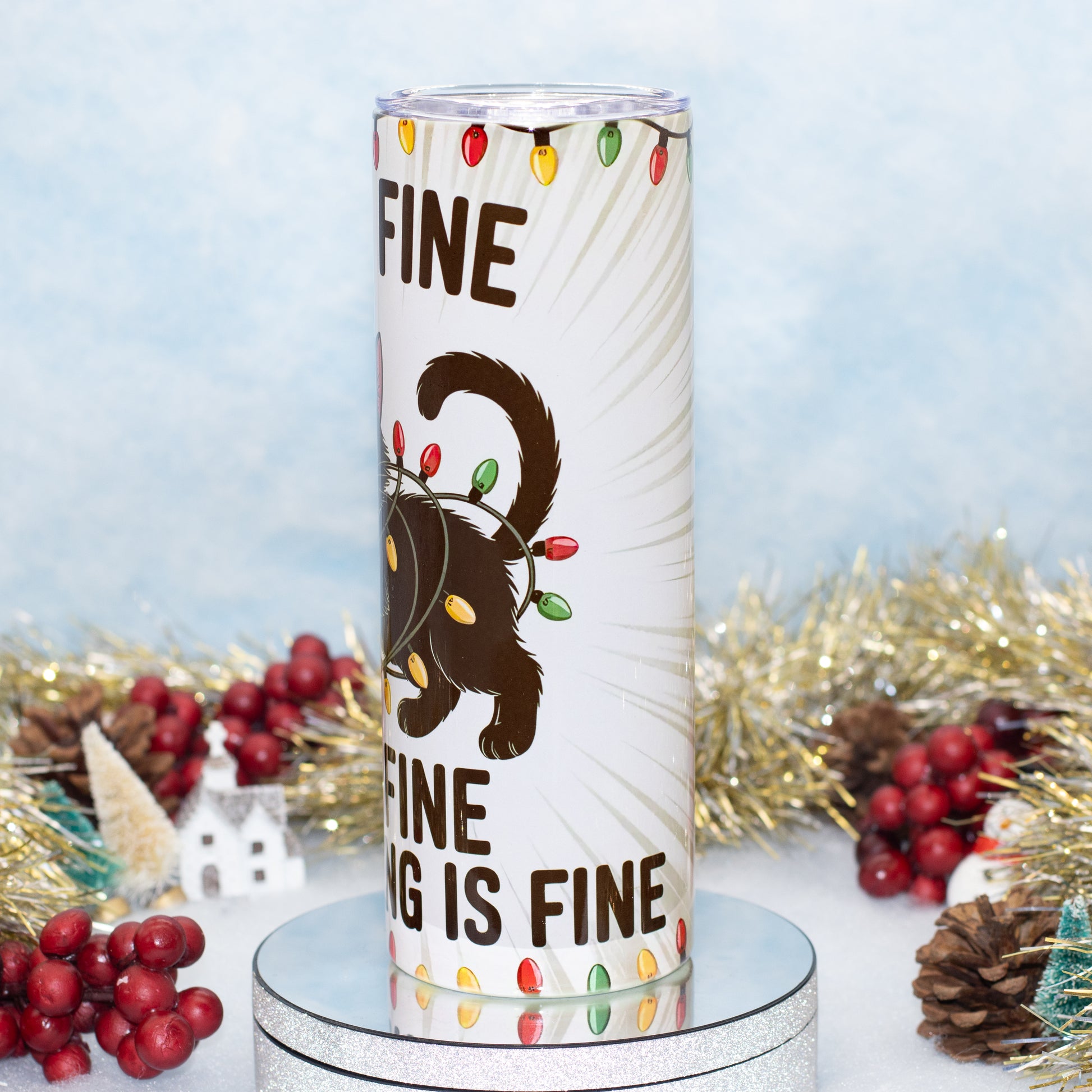 A white cylindrical tumbler with a holiday design featuring a black cat wearing a string of multicolour Christmas lights, surrounded by festive tinsel, pinecones, and red berries on a light blue background.