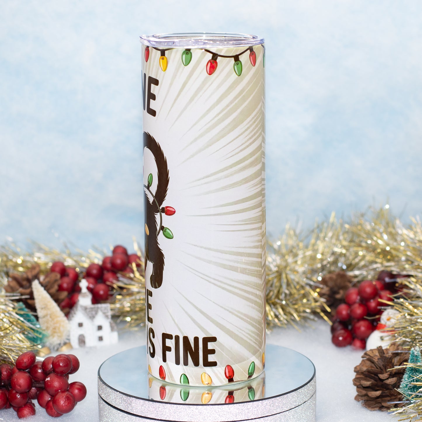 A tall white tumbler with a Christmas-themed design decorated with colourful holiday lights, displayed on a silver pedestal with festive tinsel and red berries in the background.