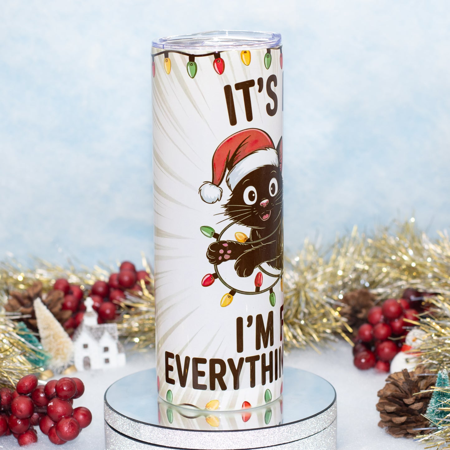 A tall white insulated tumbler with a clear lid, featuring a cartoon black cat in a red Santa hat and the text 'IT'S FINE I'M FINE EVERYTHING IS FINE' with holiday lights, displayed on a silver pedestal with Christmas decorations in the background.