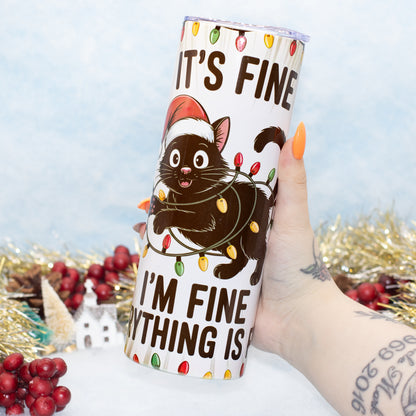 A hand holds a white insulated tumbler with a black cat illustration wrapped in Christmas lights and the text 'IT'S FINE I'M FINE. The background is a festive holiday scene with gold tinsel, red berries, and a light blue backdrop.