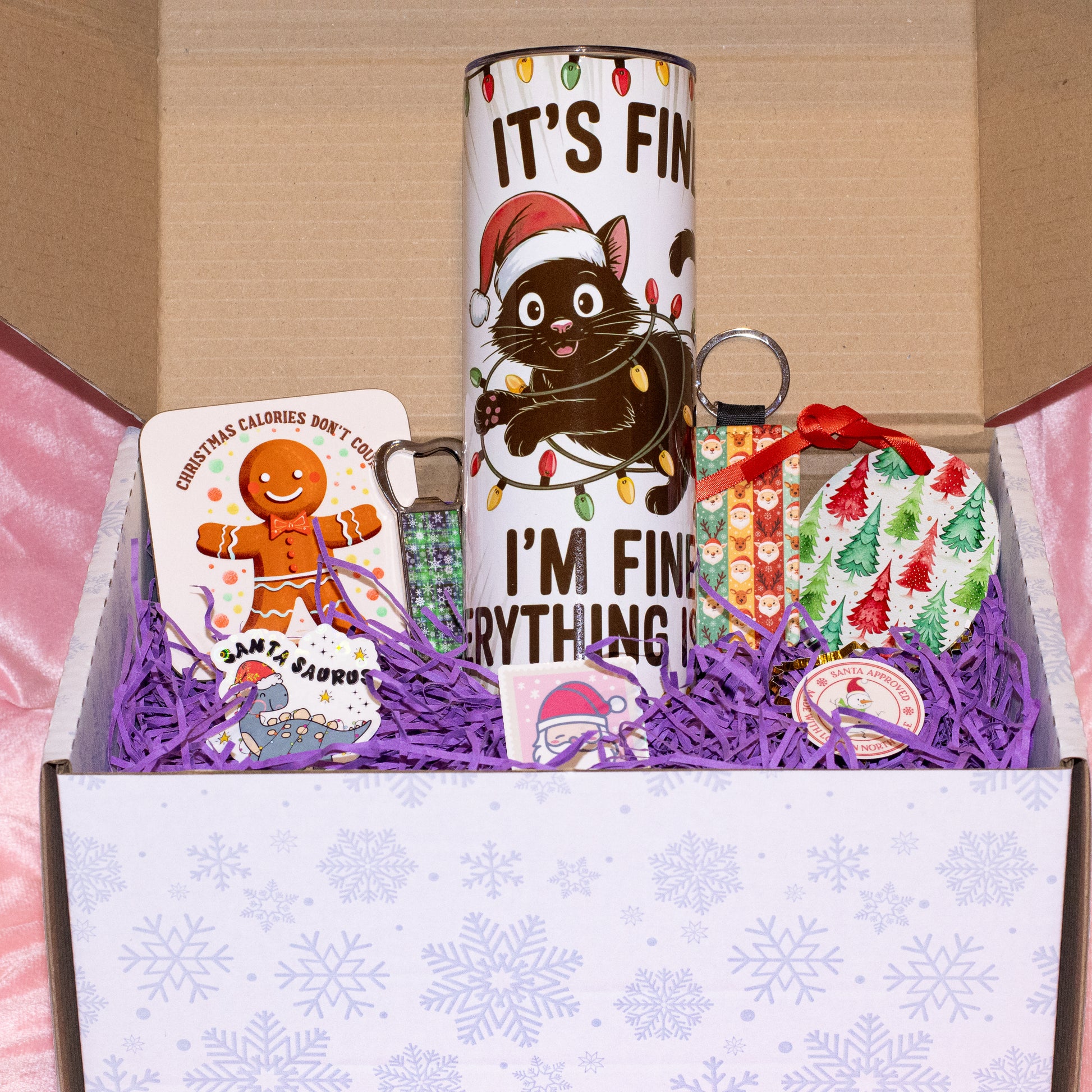 A Christmas-themed gift box set containing a white tumbler with a cartoon cat wearing a Santa hat and the text 'IT'S FIN' and 'I'M FIN', a Santa Claus card, a green beaded keychain, and a wrapped ornament, arranged in purple shredded paper inside a white box with snowflake patterns.