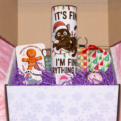 A Christmas-themed gift box set containing a white tumbler with a cartoon cat wearing a Santa hat and the text 'IT'S FIN' and 'I'M FIN', a Santa Claus card, a green beaded keychain, and a wrapped ornament, arranged in purple shredded paper inside a white box with snowflake patterns.