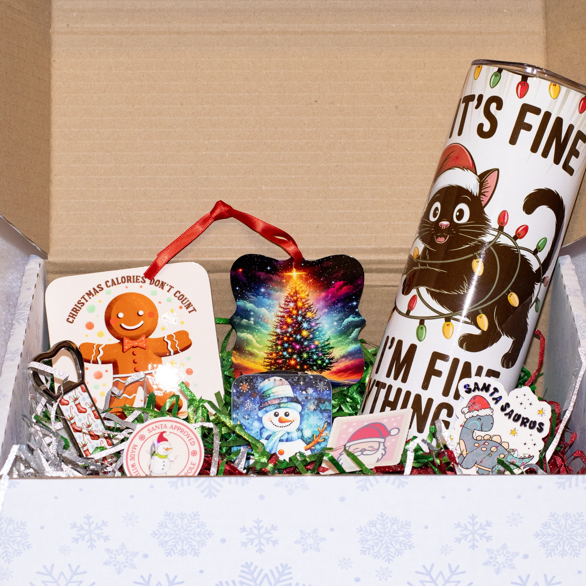 A curated Christmas gift box filled with holiday-themed items, including a gingerbread ornament card, a colorful Christmas tree ornament, a black cat mug with Christmas lights and the text 'IT'S FINE I'M NOT SICK', a Santa Sabors cookie, and a small snowman ornament, all arranged in green paper shred inside a brown cardboard box.
