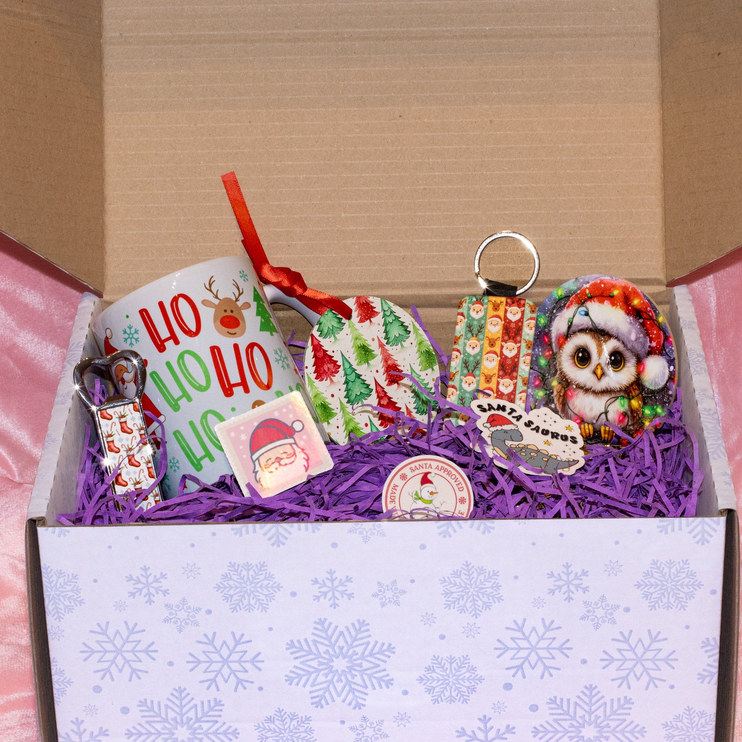 A curated Christmas gift box filled with holiday-themed items, including a white mug with 'Ho Ho Ho' and reindeer graphics, a patterned ornament, a Santa hat keychain, a owl plush or patch, and small wrapped gifts, arranged in purple shredded paper inside an open box.