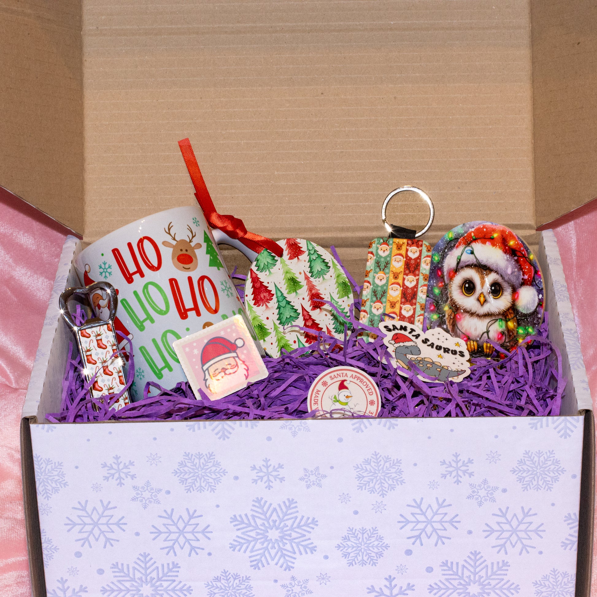 A curated Christmas gift box filled with holiday-themed items, including a white mug with 'Ho Ho Ho' and reindeer graphics, a patterned ornament, a Santa hat keychain, a owl plush or patch, and small wrapped gifts, arranged in purple shredded paper inside an open box.