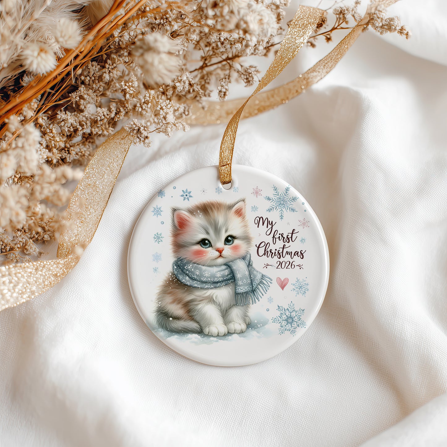 A round ceramic Christmas ornament featuring an illustration of a grey and white kitten wearing a blue scarf, with the text 'My first Christmas 2026' and snowflake designs. The ornament hangs from a gold ribbon on a soft white fabric background with dried flowers and a gold ribbon.