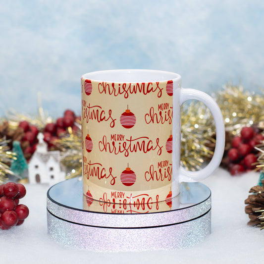 A white ceramic mug with a beige printed wrap featuring 'Merry Christmas' in red script and red ornament illustrations, displayed on a round silver glitter pedestal with holiday decorations in the background.