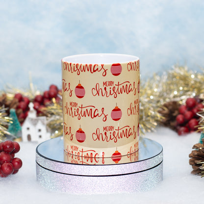 A ceramic mug with a festive Christmas print featuring 'Merry Christmas' in red script and red ornament illustrations on a cream background, displayed on a silver glitter pedestal with holiday decorations in the background.