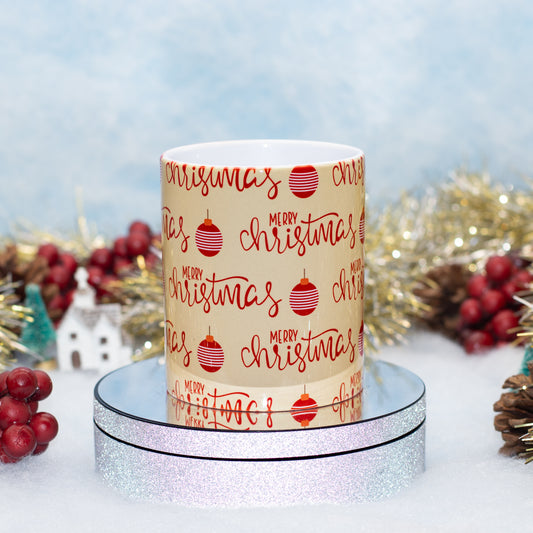 A ceramic mug with a festive Christmas print featuring 'Merry Christmas' in red script and red ornament illustrations on a cream background, displayed on a silver glitter pedestal with holiday decorations in the background.