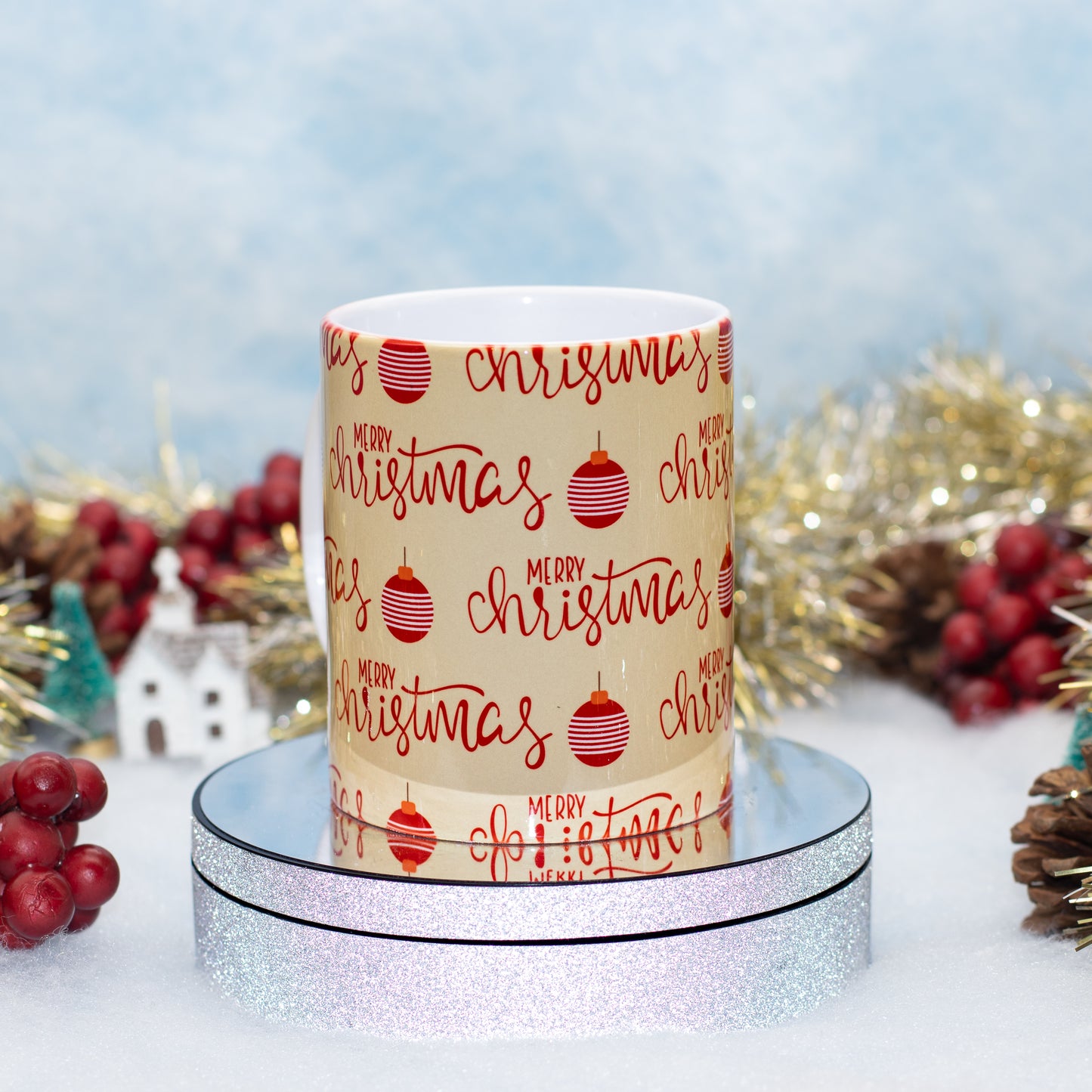 A white ceramic mug wrapped in a beige holiday paper print with red 'Merry Christmas' script and red ornament motifs, displayed on a round silver glitter stand with festive tinsel and berries in the background.