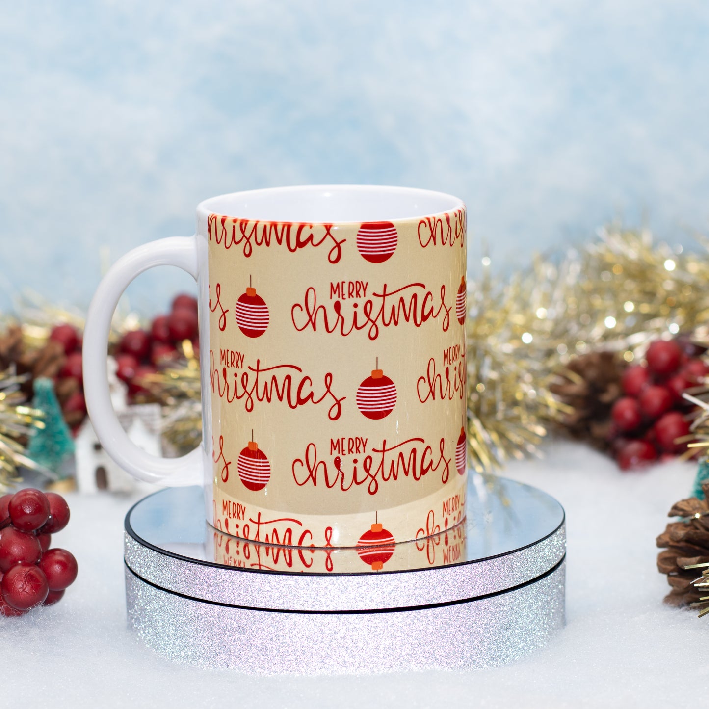 A white ceramic mug with a beige printed wrap featuring red 'Merry Christmas' script and striped ornament illustrations, displayed on a round silver glitter pedestal with festive tinsel and berries in the background.