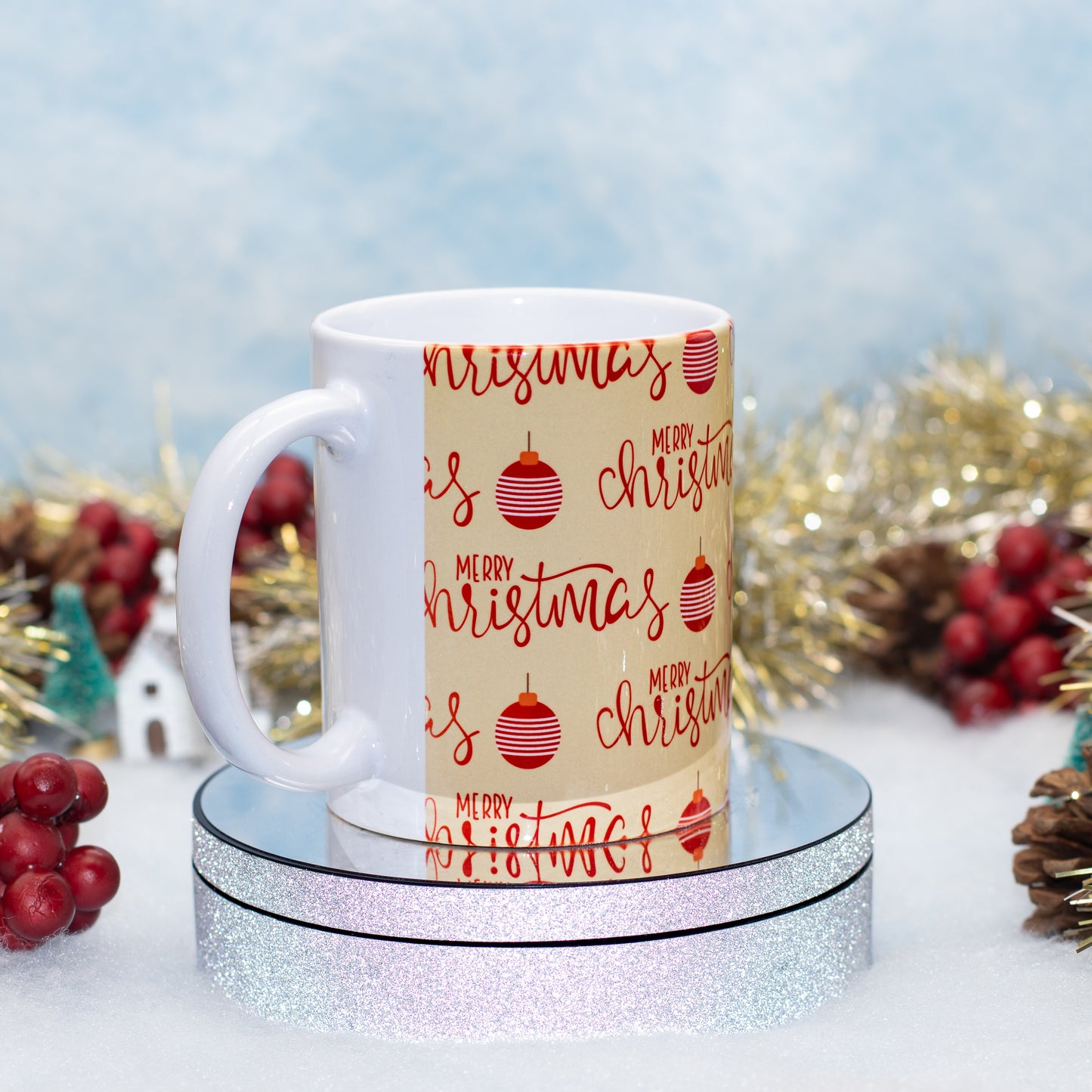 A white ceramic mug with a beige holiday wrap design featuring red 'Merry Christmas' script and ornaments, displayed on a round silver glitter pedestal with festive blurred background decorations.