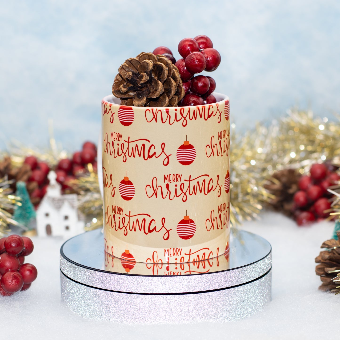 A ceramic mug wrapped in festive Christmas paper with 'Merry Christmas' and red ornaments, topped with a pinecone and red berries, displayed on a round silver glitter pedestal.
