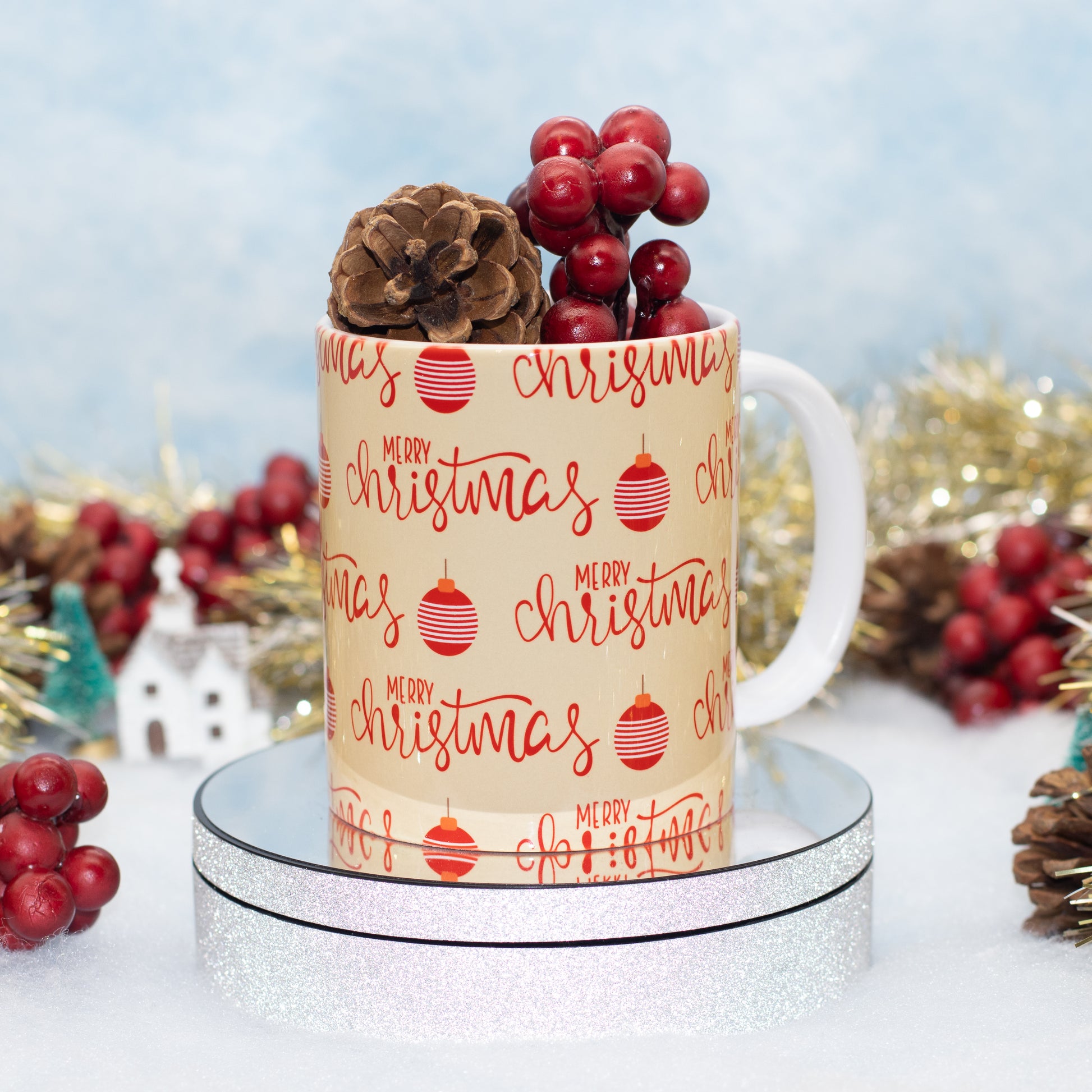A white ceramic mug with a beige printed pattern reading 'Merry Christmas' in red script and red ornament illustrations, styled with red berries and a pinecone on a reflective silver stand against a soft holiday backdrop.