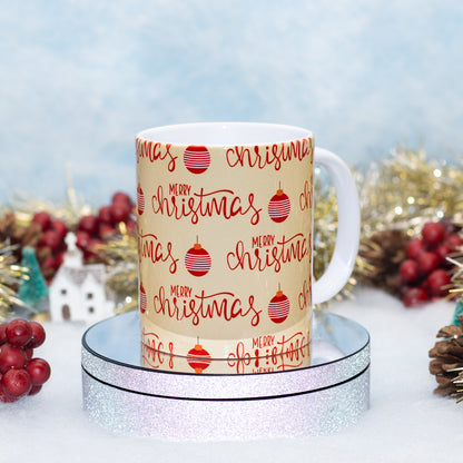 A white ceramic mug with a beige printed wrap featuring the phrase 'Merry Christmas' in red script and red ornament illustrations, displayed on a silver glitter ring with festive holiday decorations in the background.
