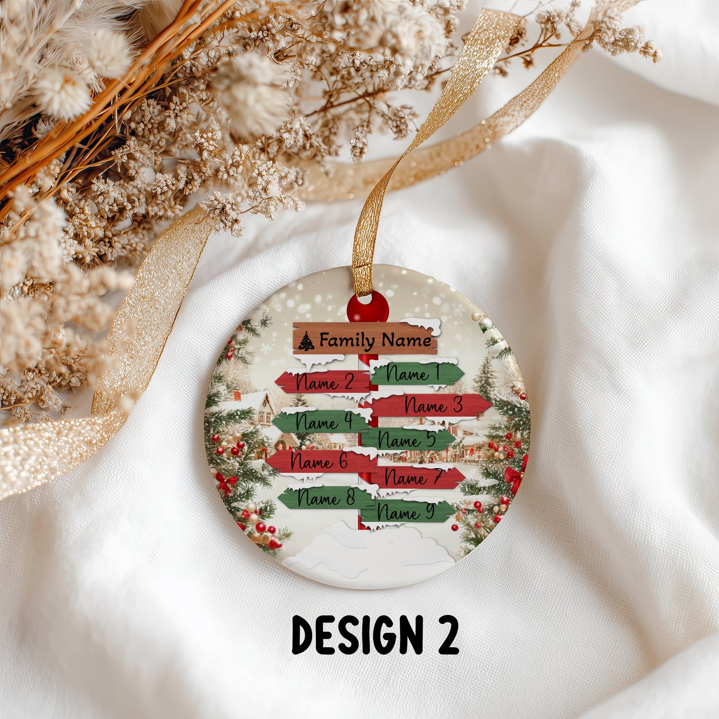 A round Christmas ornament with a personalized direction-sign design, hanging from a gold ribbon on a white fabric background with dried flowers. The ornament features red, green, and brown sign elements with placeholder text and the label 'DESIGN 2' below.