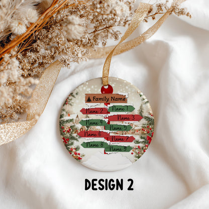 A round Christmas ornament with a personalized direction-sign design, hanging from a gold ribbon on a white fabric background with dried flowers. The ornament features red, green, and brown sign elements with placeholder text and the label 'DESIGN 2' below.