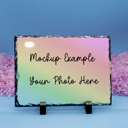 A rectangular slate photo plaque on a small black stand, featuring a pastel gradient background with the text 'Mockup Example Your Photo Here' in black script. The plaque is photographed against a blue backdrop with pink shredded paper on both sides.